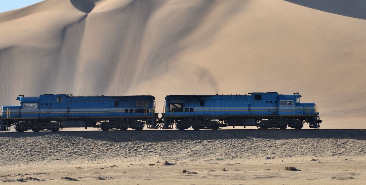 Two diesel locomotives running beneath a high sand dune